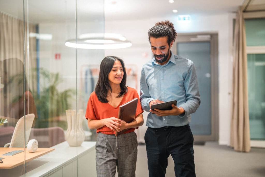 istockphoto-1487231759-2048×2048 Two handsome, diverse colleagues are seen engaging in a productive conversation while utilizing wireless technology and a digital tablet in a modern office setting. Their smart casual attire adds to their professional appearance as successful employees.